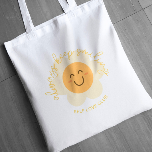 White tote bag with a happy flower face design and text on a wooden surface