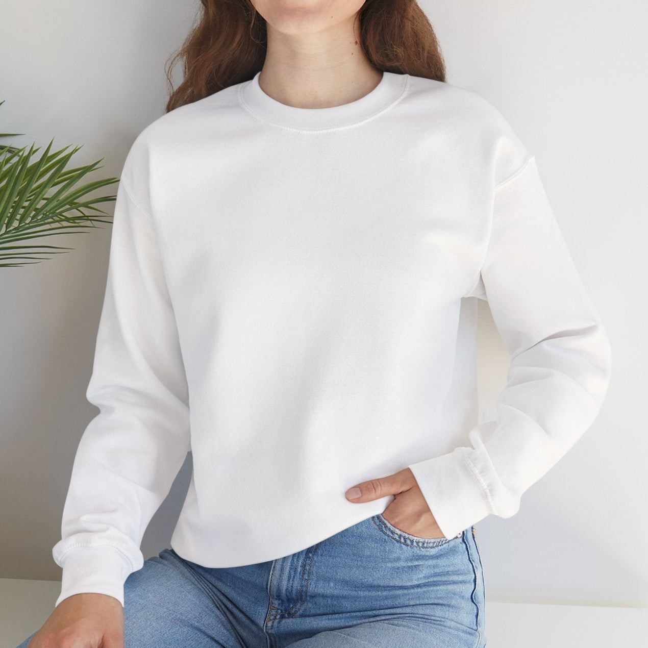 Woman wearing a white sweatshirt and blue jeans sitting on a white surface with a plant in the background.