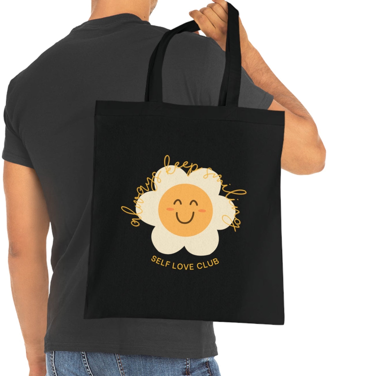 Person holding a black tote bag with a smiling face flower design and 'Self Love Club' text.