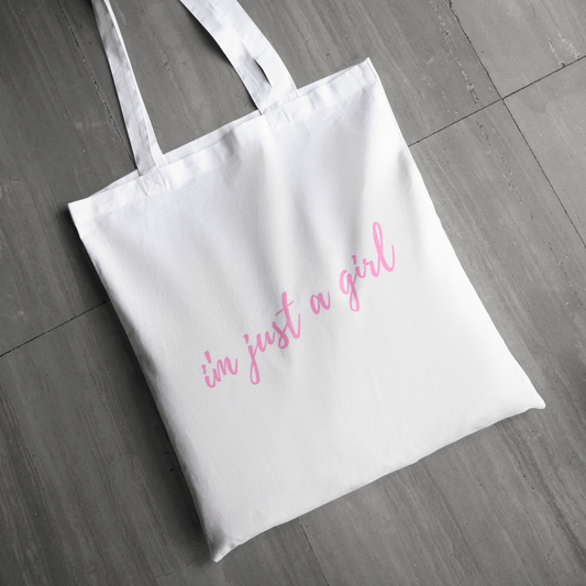 White tote bag with 'I'm just a girl' text in pink on a wooden floor.