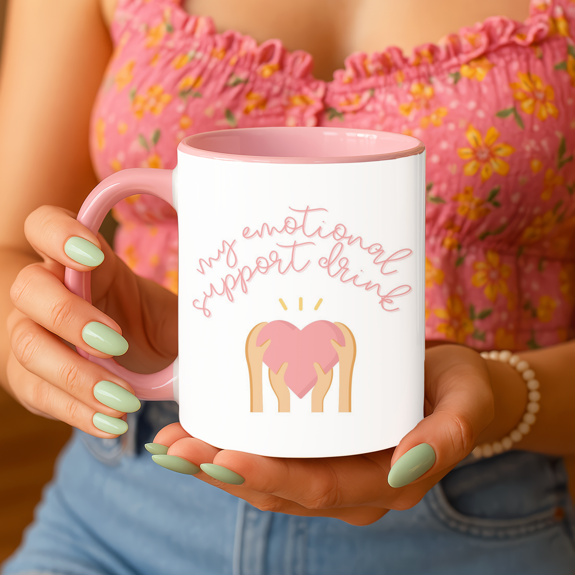 Person holding a mug with a heart design and text, wearing a floral top and jeans.